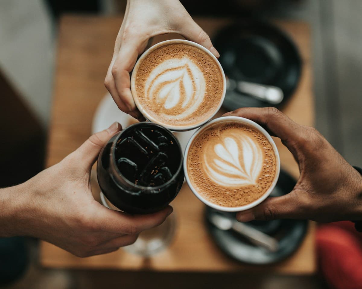 Tres personas brindan con dos cafés con leche y un café frío sobre una mesa de madera